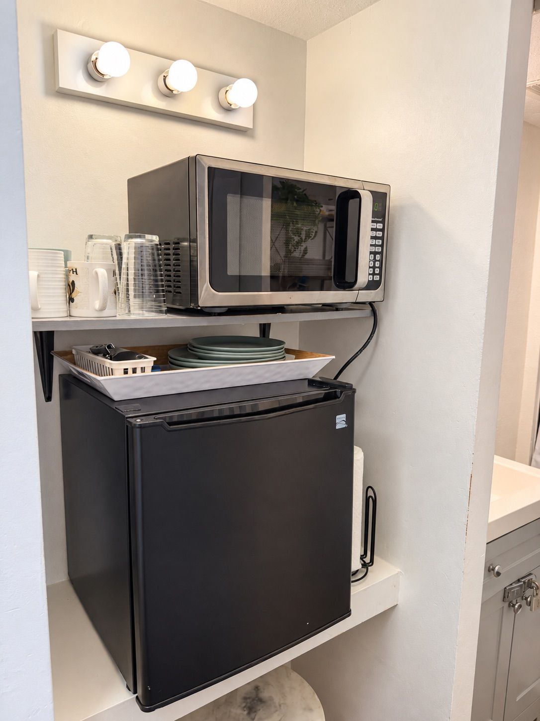Une petite cuisine équipée d’un four à micro-ondes et d’un petit réfrigérateur, une étagère avec des assiettes et des tasses empilées, et un luminaire fixé au mur.