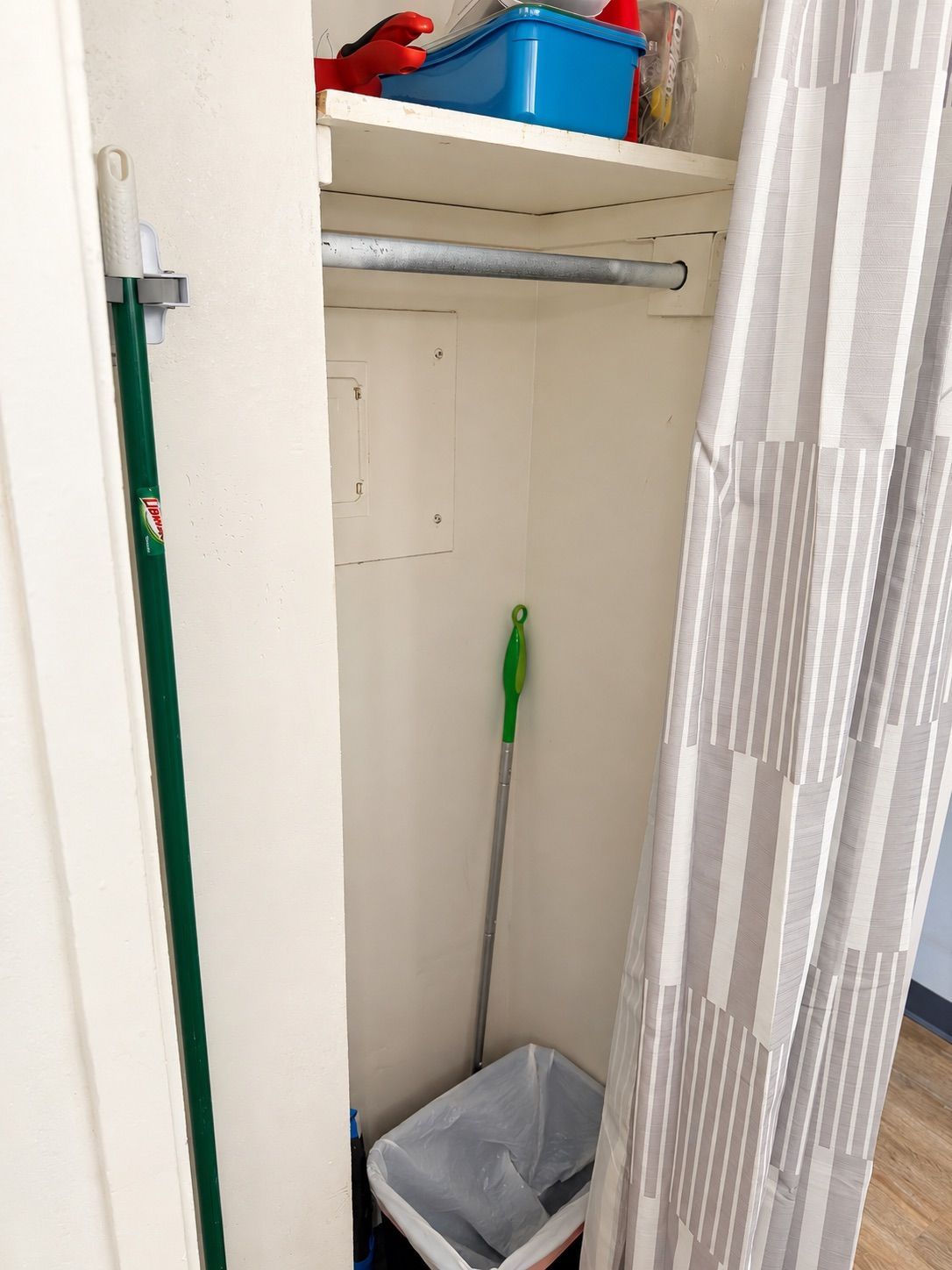 Un baño estrecho con ducha, un trapeador verde, un bote de basura blanco y un caja azul en la pared.