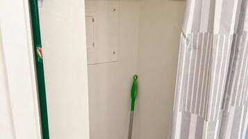 Un baño estrecho con ducha, un trapeador verde, un bote de basura blanco y un caja azul en la pared.