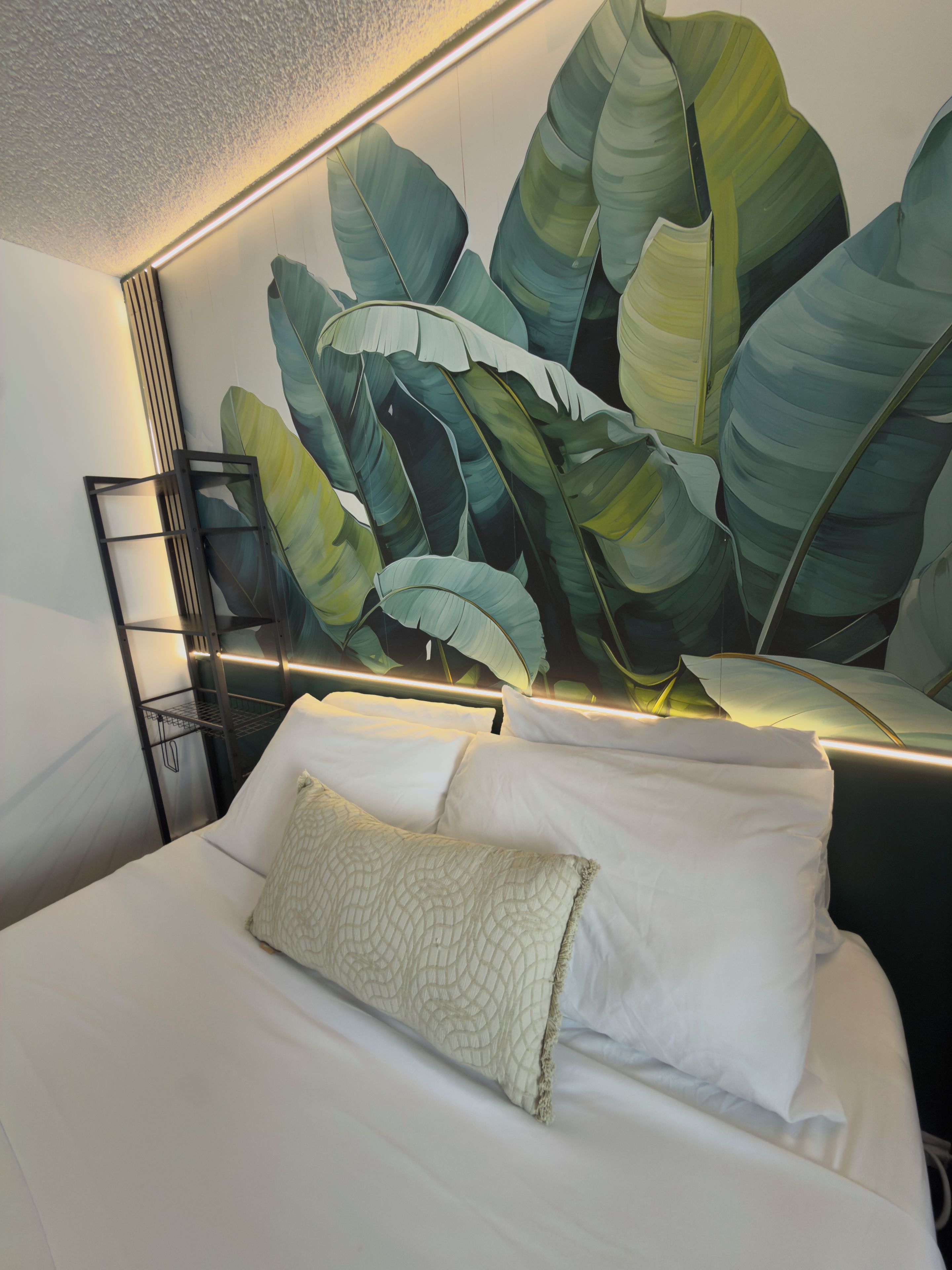 A hotel room with a large bed, white bedding, a headboard with a leaf pattern, and a black metal shelf unit.
