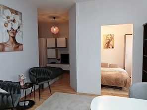 A modern living room with a black leather sofa, a small side table, and a white armchair. There is a painting on the wall and a ceiling light fixture.