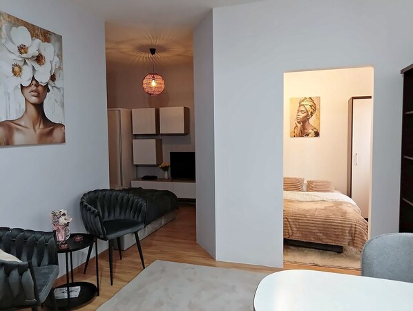 A modern living room with a black leather sofa, a small side table, and a white armchair. There is a painting on the wall and a ceiling light fixture.