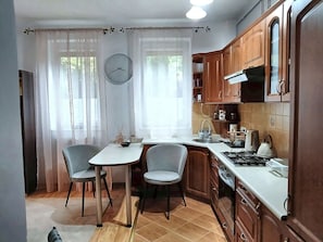 A compact kitchen with a dining table, two chairs, a clock, and wooden cabinets.