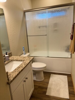 Une salle de bain avec un lavabo sur un plan de travail en granit, des toilettes, une douche en verre et un tapis blanc sur le sol.