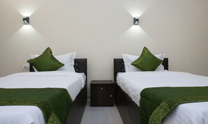 Quarto de hotel com duas camas, cada uma com manta verde, roupa de cama branca e almofadas verdes a combinar. Há mesas de cabeceira com luminárias e uma pequena mesinha de cabeceira entre as camas.