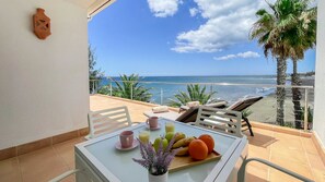 A balcony with a table set for breakfast, overlooking a beach with palm trees and the ocean. - Nueva Suecia 716 Studio (San Agustin)