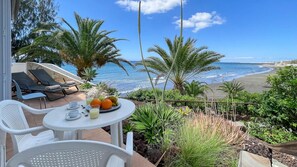 A balcony with a table set for two, overlooking a beach with palm trees and ocean waves. - Nueva Suecia 716 Studio (San Agustin)