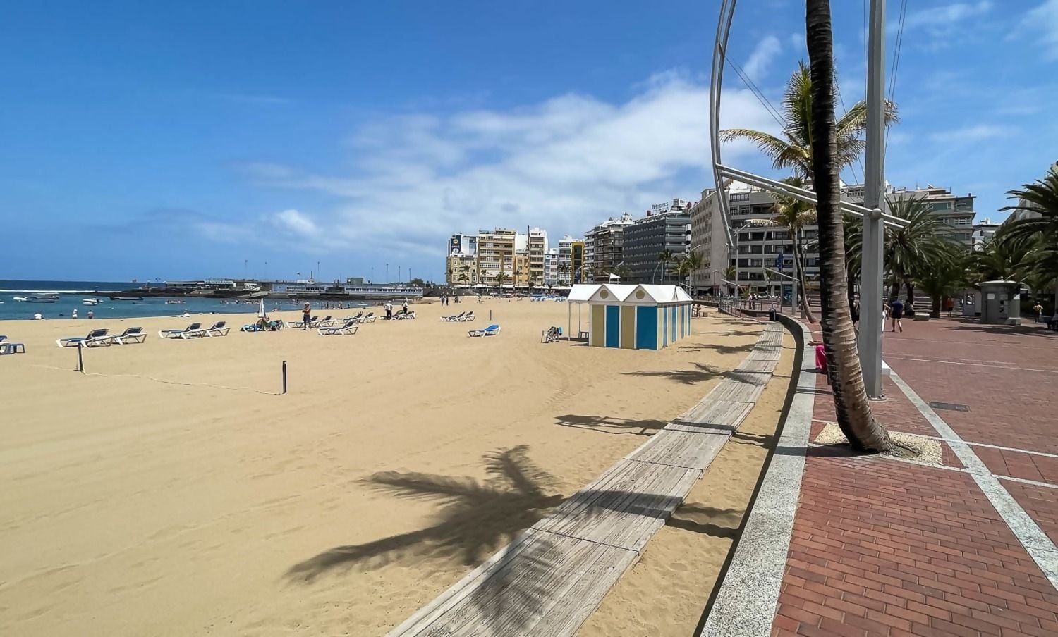 A beach with sun loungers, a wooden boardwalk, and a row of buildings in the background.