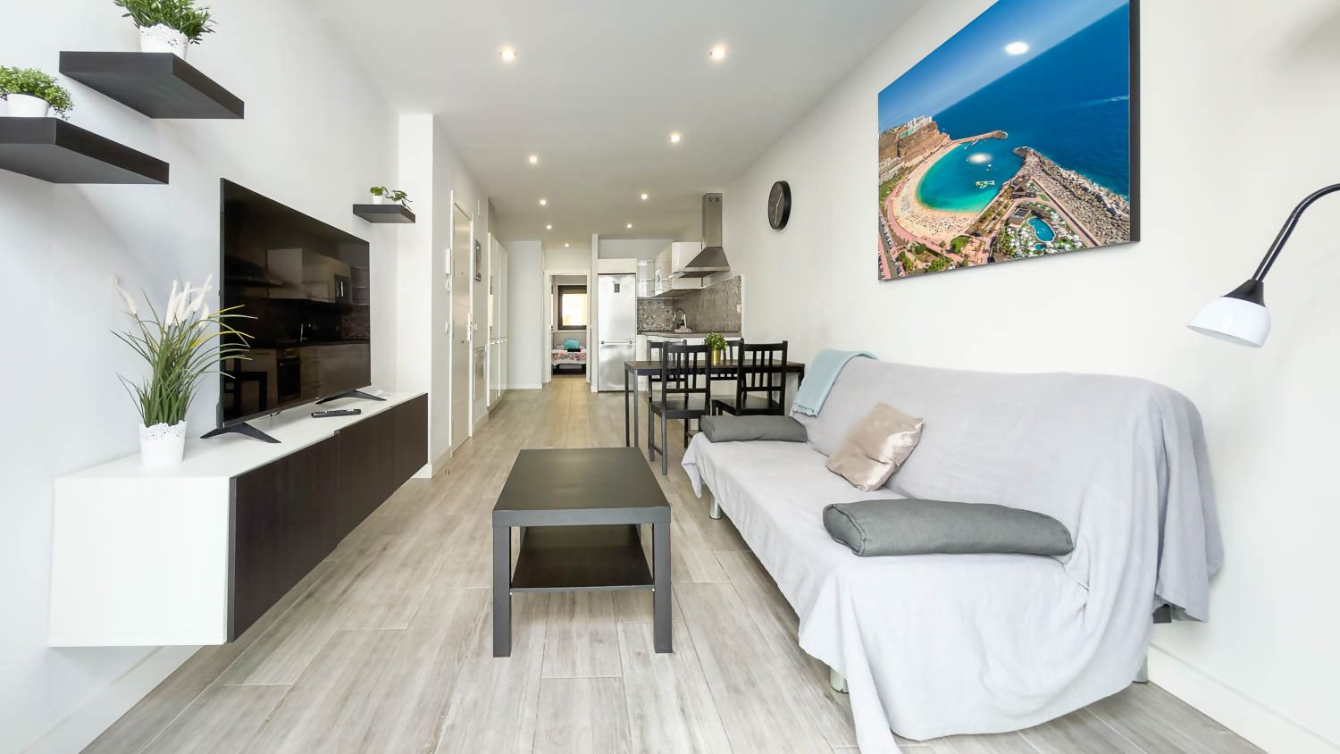 Een moderne woonkamer met een bank, een salontafel en een televisie. Er is een eethoek met een tafel en stoelen. Op de muur hangt een schilderij van een strand.