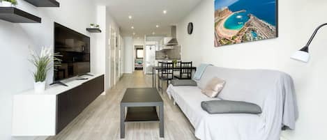 A modern living room with a sofa, a coffee table, and a TV. There is a dining area with a table and chairs. A painting of a beach is on the wall.