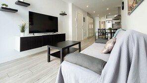 Un salon moderne avec un téléviseur à écran plat, un meuble de rangement en bois foncé, un canapé gris avec des coussins et, au fond, un coin repas.