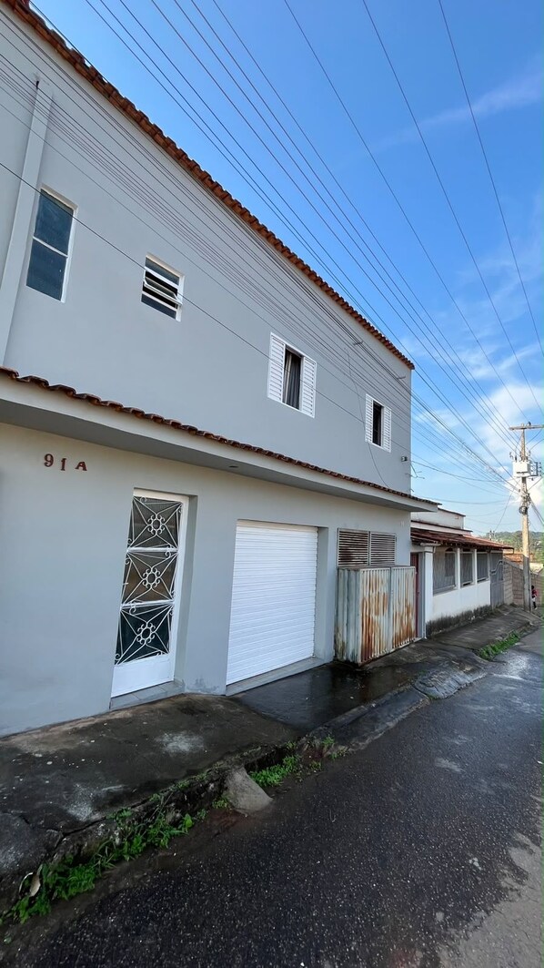 Un edificio de dos pisos con fachada blanca, número 91A, que cuenta con una puerta de garaje, un ventanal con contraventanas y un portón de metal.