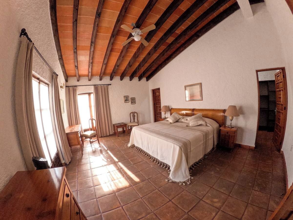 Une chambre à coucher avec un grand lit, des poutres en bois au plafond, une table de chevet avec une lampe et une fenêtre avec des rideaux.