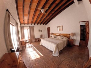 Une chambre à coucher avec un grand lit, des poutres en bois au plafond, une table de chevet avec une lampe et une fenêtre avec des rideaux.