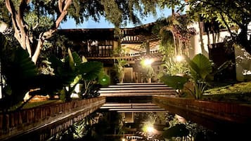 Une structure en bois à plusieurs niveaux, dotée d’un escalier central, entourée d’une végétation luxuriante et agrémentée d’un élément aquatique décoratif.
