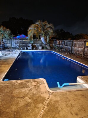 En rektangulær swimmingpool med blå overflade, omgivet af en betonflisebelagt terrasse og et træhegn.