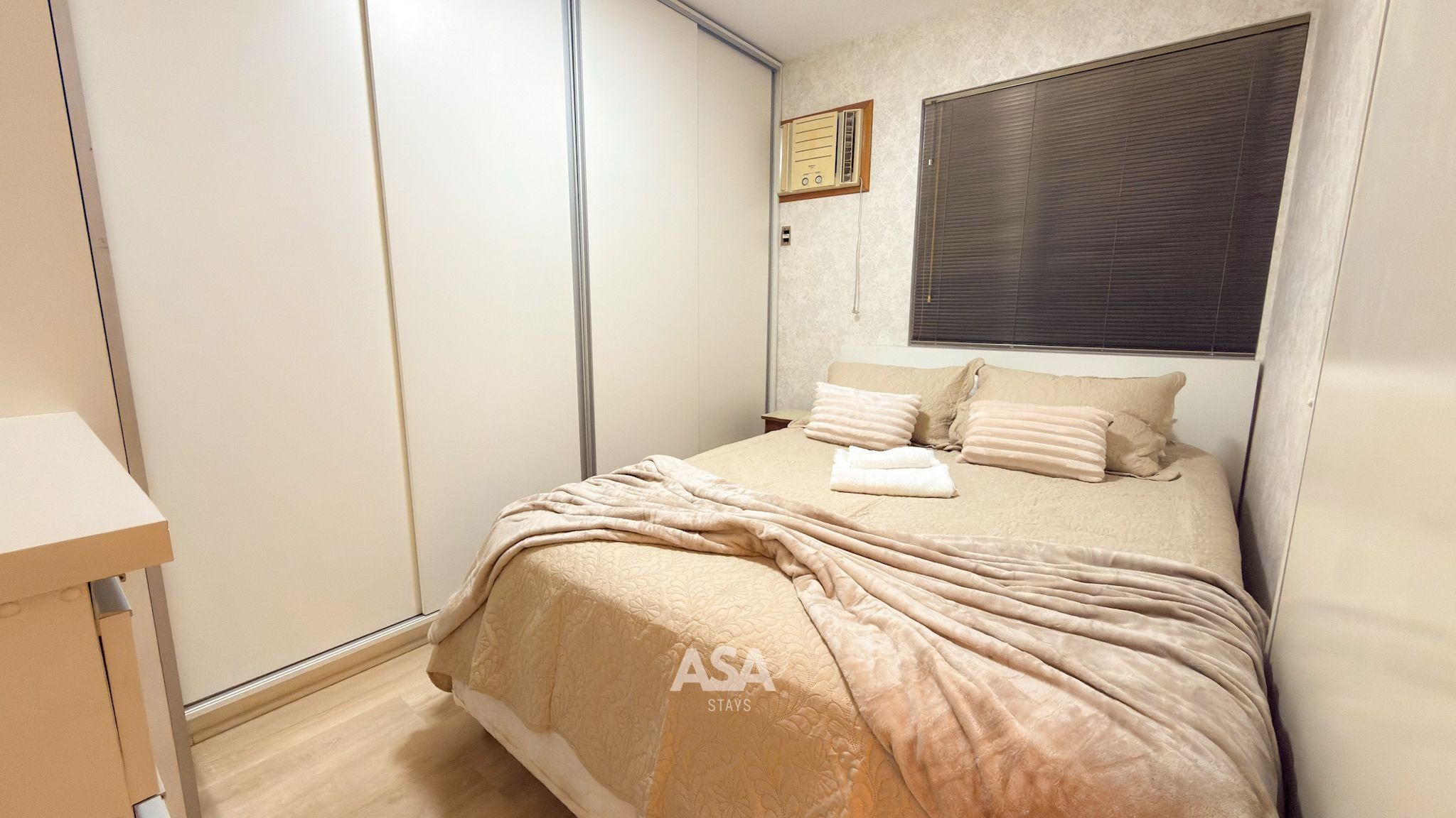 Ein Schlafzimmer mit einem Bett, Kissen, einem Nachttisch, einem Kleiderschrank und einem Fenster mit Jalousien.