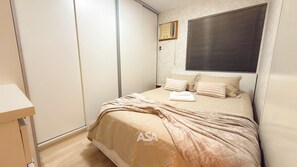 Un dormitorio con cama, almohadas, mesita de noche, armario y ventana con persianas.