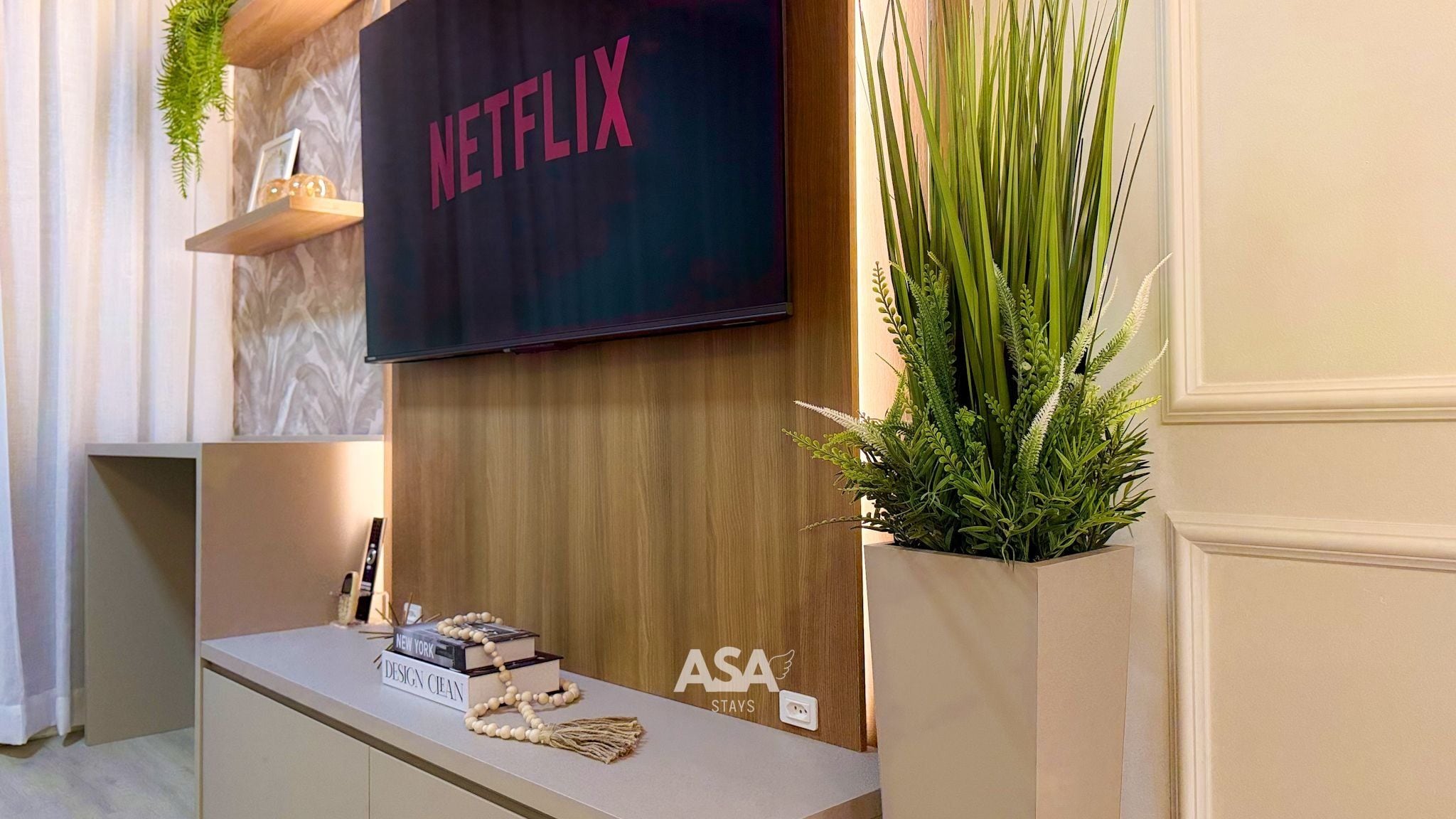 A room with a flat-screen TV mounted on a wall, a wooden console table with books and decorative items, and a potted plant.