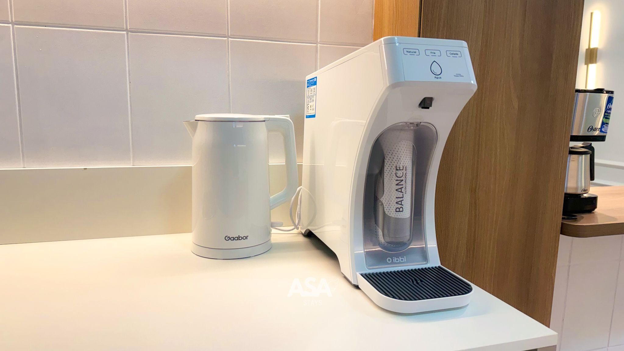 Une cuisine moderne équipée d’une machine à café, d’une bouilloire et d’un distributeur d’eau, le tout posé sur un plan de travail blanc.
