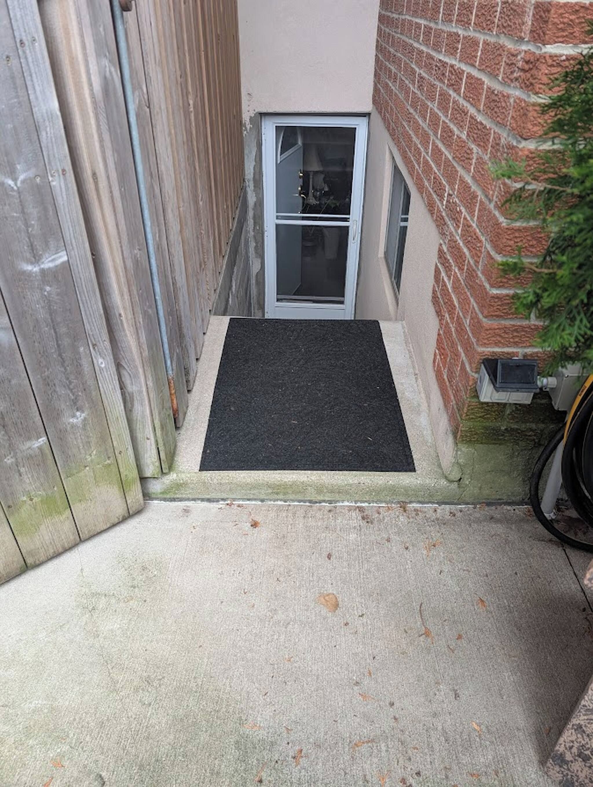 A narrow outdoor corridor with a wooden fence on one side and a brick wall on the other. There is a small door with a glass panel and a black mat at the entrance.