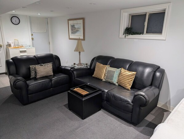A living room with a black leather sofa, a coffee table, and a side table with a lamp. There is a clock on the wall and a window with a plant on the sill. - Room-king Size In-suite Basement (Toronto)