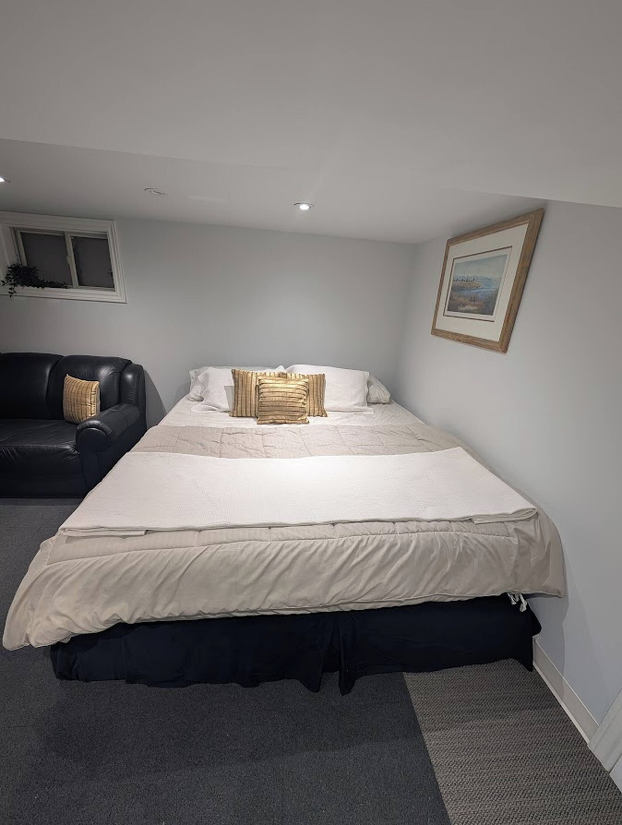 A single bed with a white mattress and a beige blanket, a black leather sofa, a framed picture on the wall, and a window with a plant.