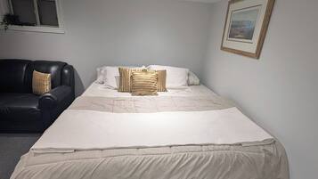 A single bed with a white mattress and a beige blanket, a black leather sofa, a framed picture on the wall, and a window with a plant.