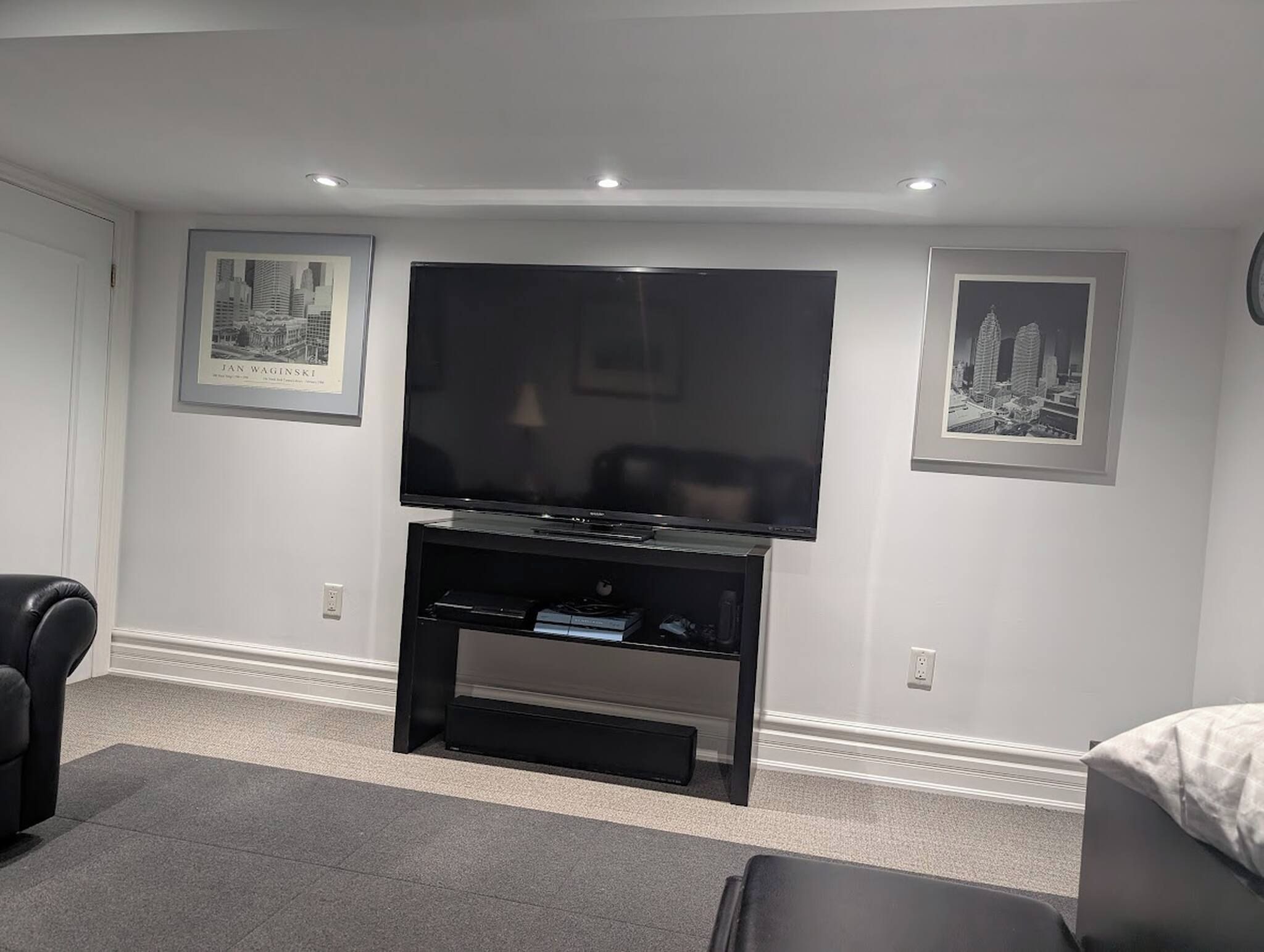 Una sala de estar con un televisor de pantalla plana sobre un soporte negro, dos cuadros con vistas a la ciudad en la pared, un sillón de cuero negro y una alfombra gris.