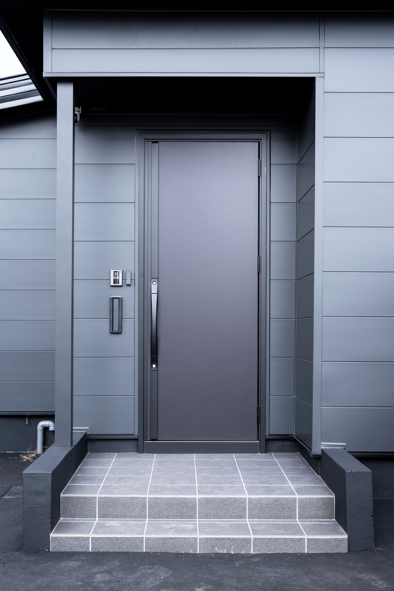 A modern building entrance with a grey door, a small step leading up to it, and a security keypad on the left.