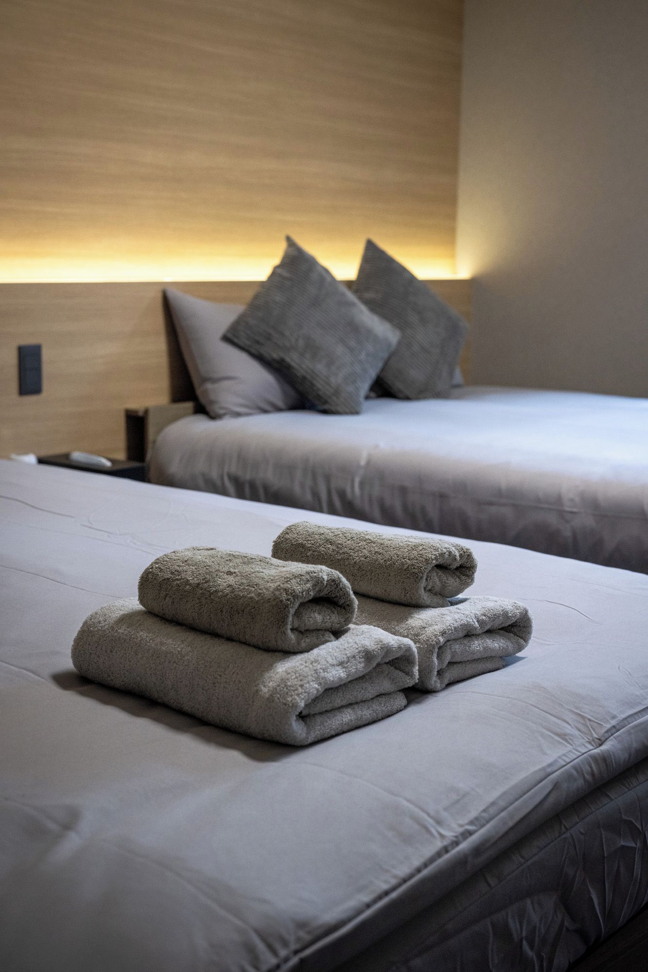 Two beds with neatly folded towels, a wooden headboard, and a wall-mounted light.