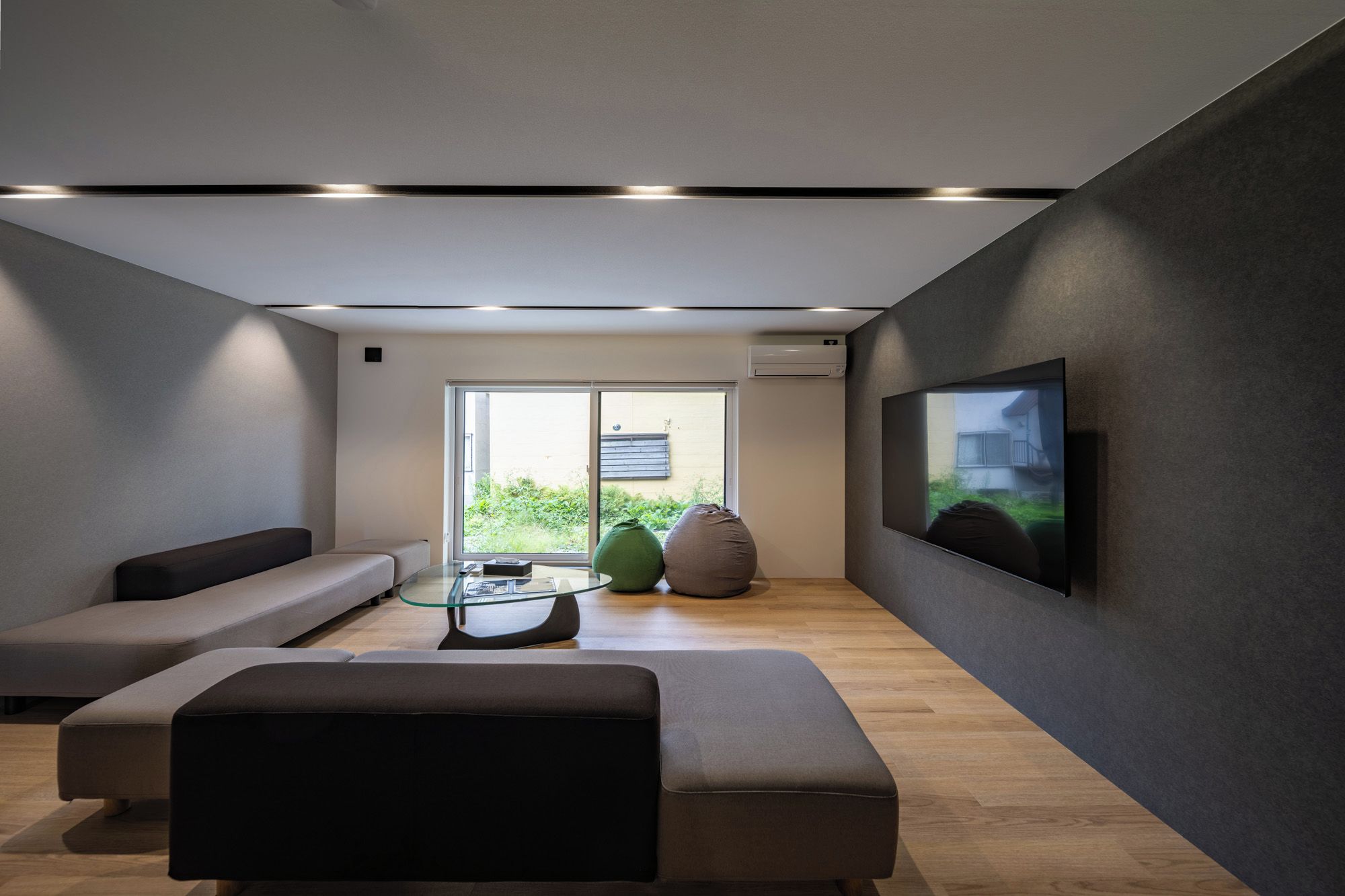 A modern living room with a glass coffee table, a sofa, a bean bag, and a flat-screen TV mounted on the wall.