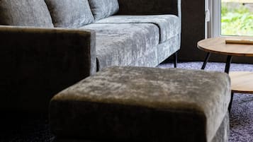 A room with a grey sofa and a matching ottoman, a small round table, and a window with a view of greenery.