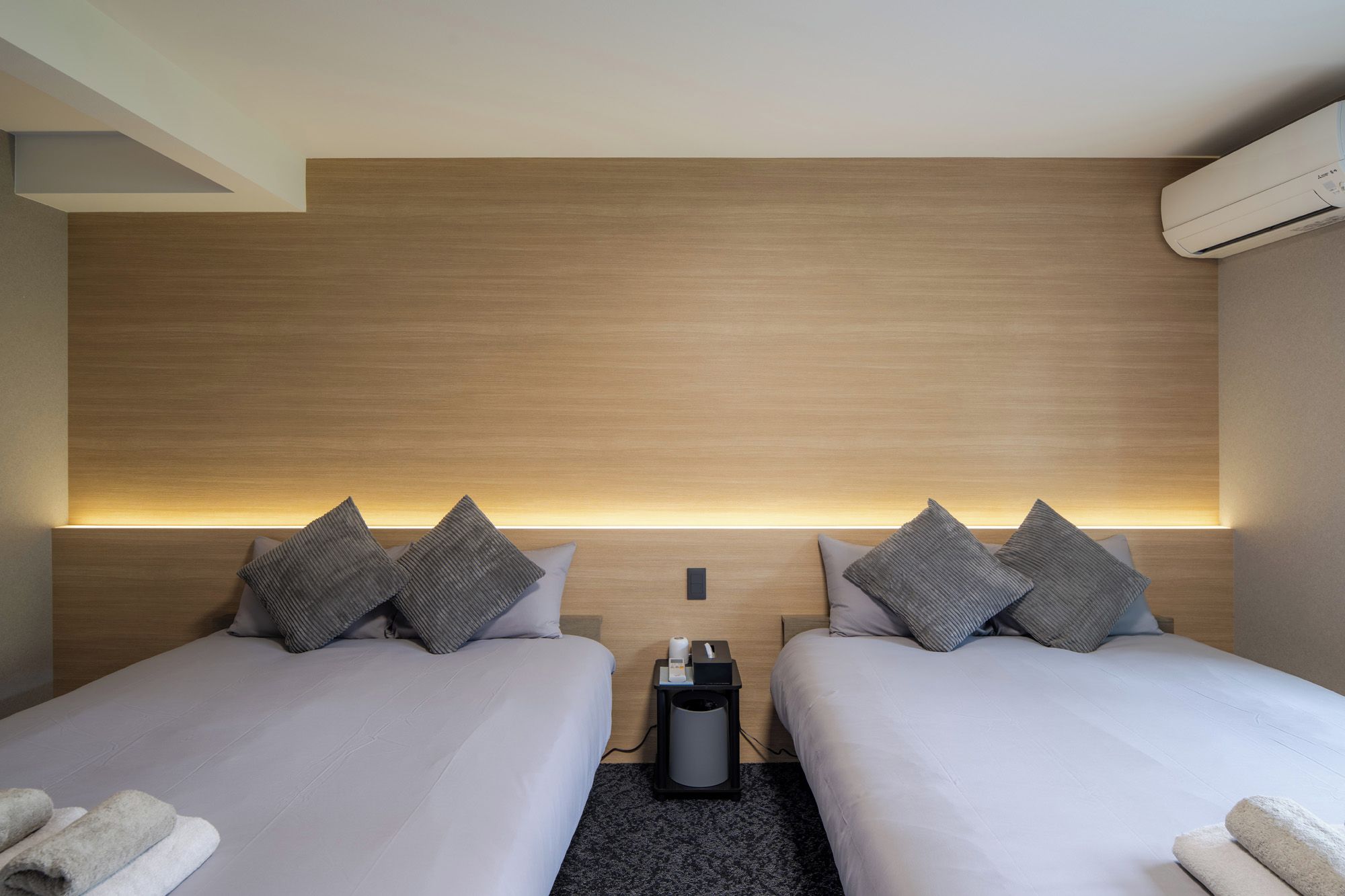 A hotel room with two beds, a nightstand, a trash bin, and an air conditioning unit.
