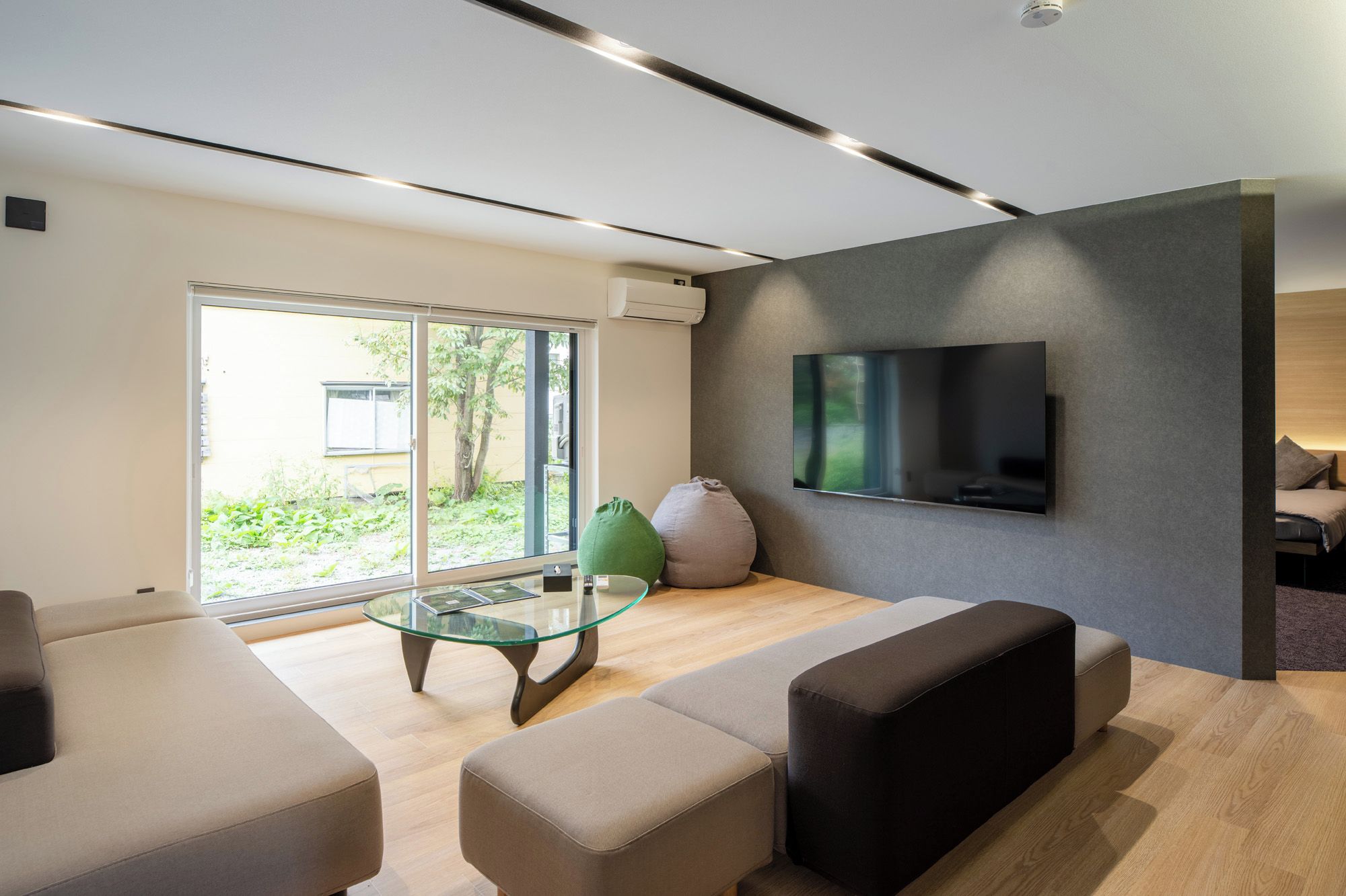 A modern living room with a glass coffee table, sofas, a flat-screen TV mounted on the wall, and a sliding glass door leading to an outdoor area.
