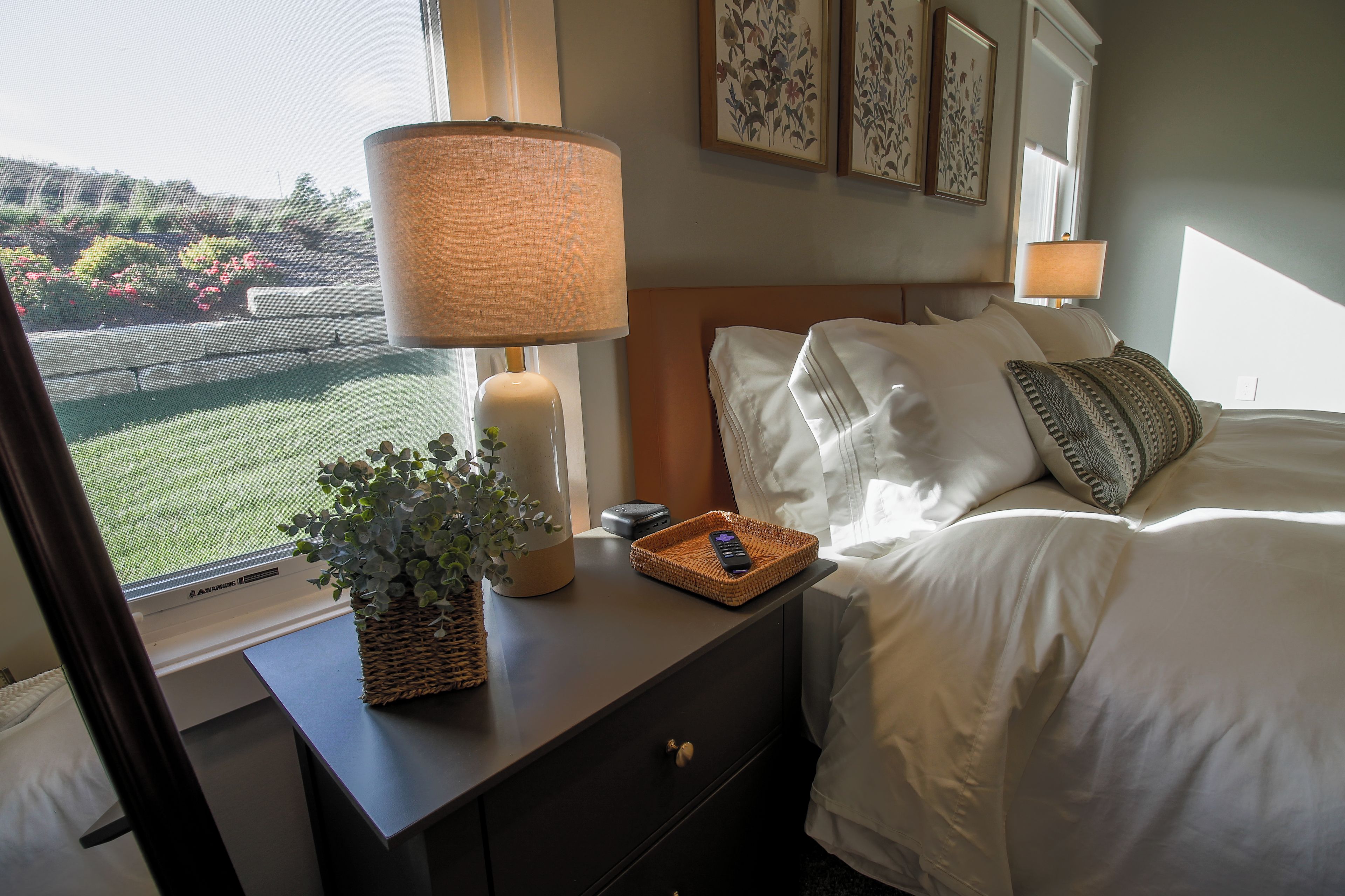 A hotel room with a bed, bedside lamps, a nightstand with a remote control, a decorative plant, and framed artwork on the wall.