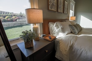 A hotel room with a bed, bedside lamps, a nightstand with a remote control, a decorative plant, and framed artwork on the wall.