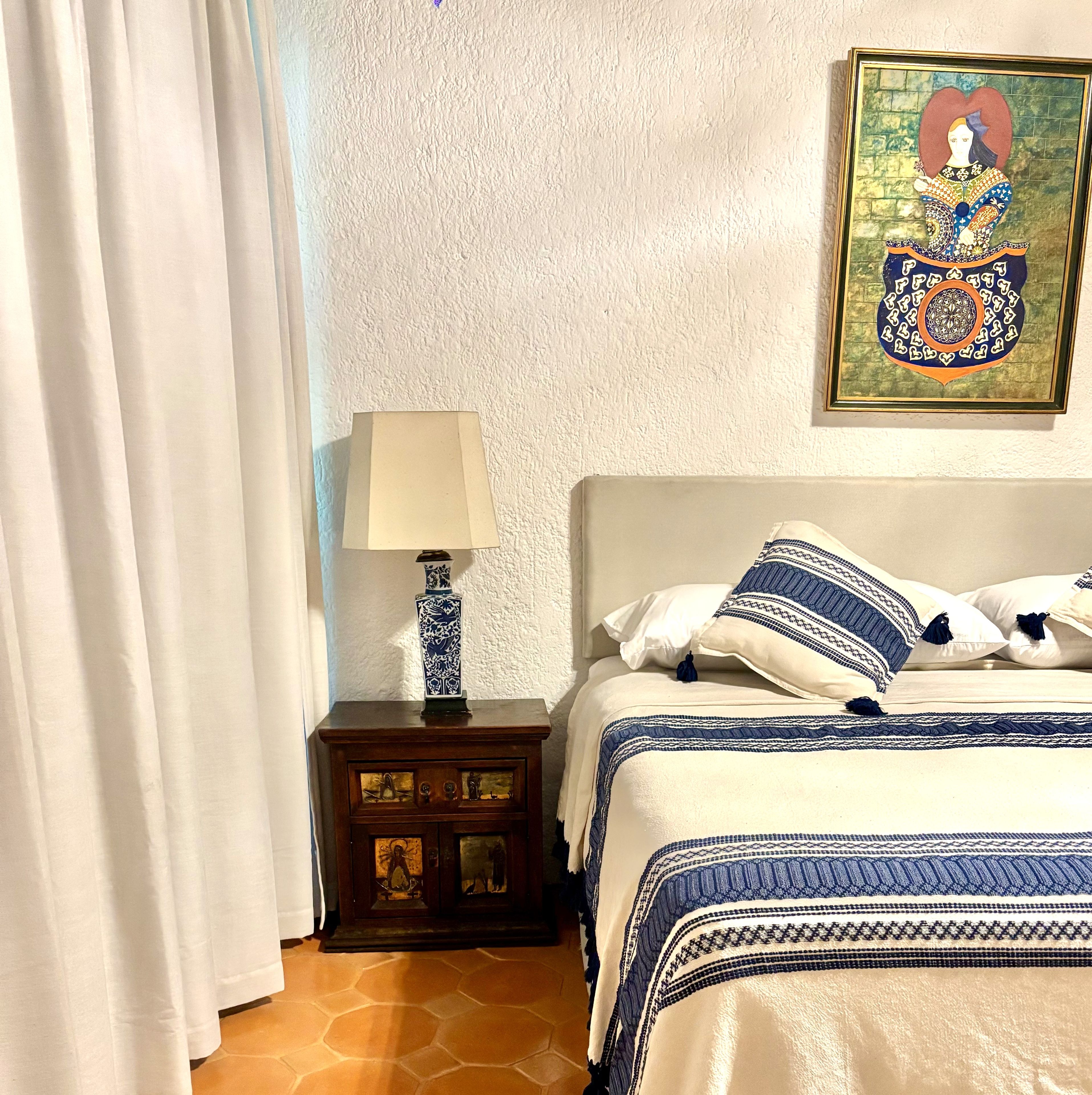 Un dormitorio con cama, una mesita de noche con lámpara, un cuadro en la pared y una ventana con cortinas.