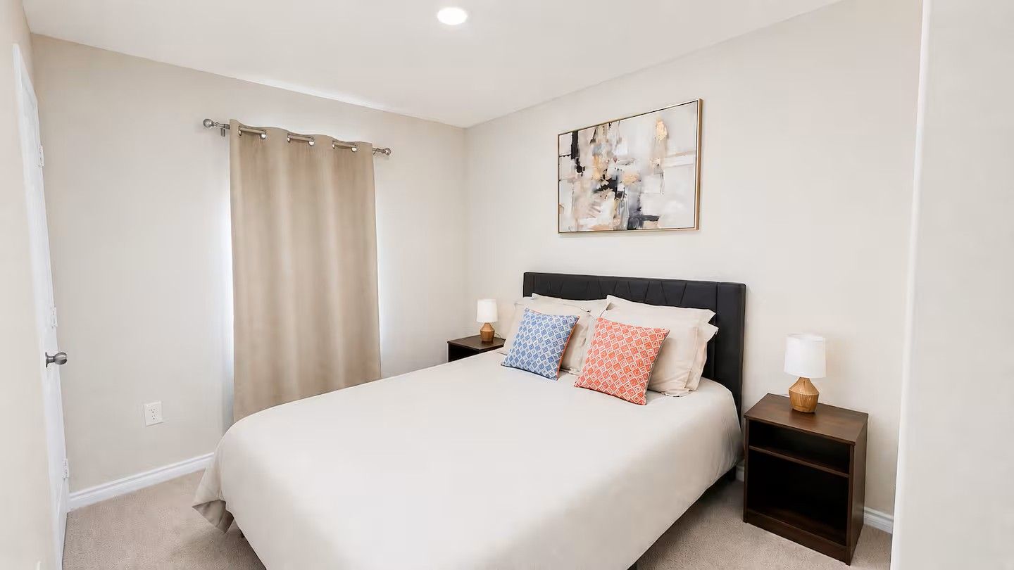A hotel room with a bed, two bedside tables, a nightstand, a curtain, and a wall-mounted abstract painting.