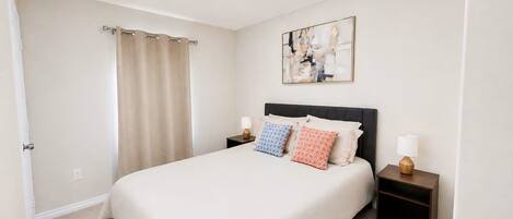 A hotel room with a bed, two bedside tables, a nightstand, a curtain, and a wall-mounted abstract painting.