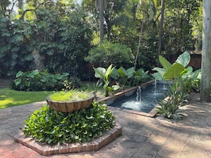 Un jardin agrémenté d’un élément aquatique, d’un bac en pierre contenant des plantes et d’un chemin pavé de briques.
