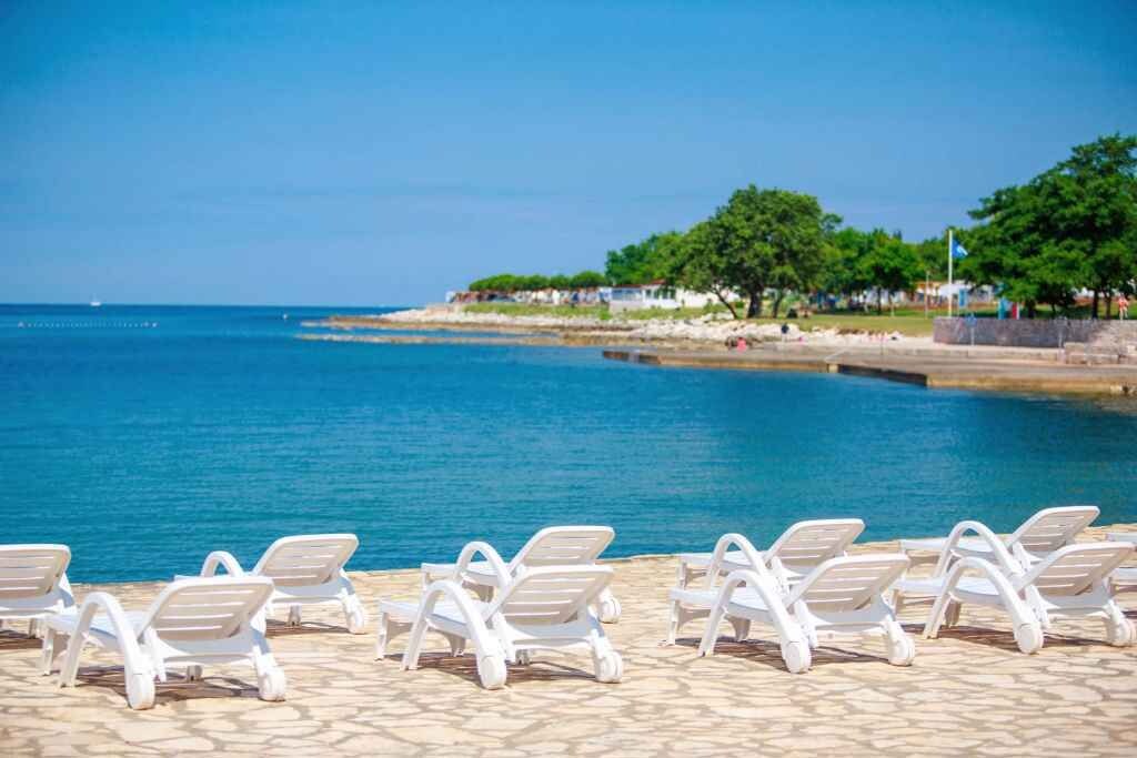 A beachside view with lounge chairs facing the sea, a clear sky, and a distant shoreline with greenery and buildings.
