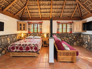 Een slaapkamer met een stenen muur, een houten bed, een bankje en een rieten dak.