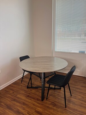 Une pièce avec une table ronde et quatre chaises, une fenêtre avec des stores et un plancher en bois.