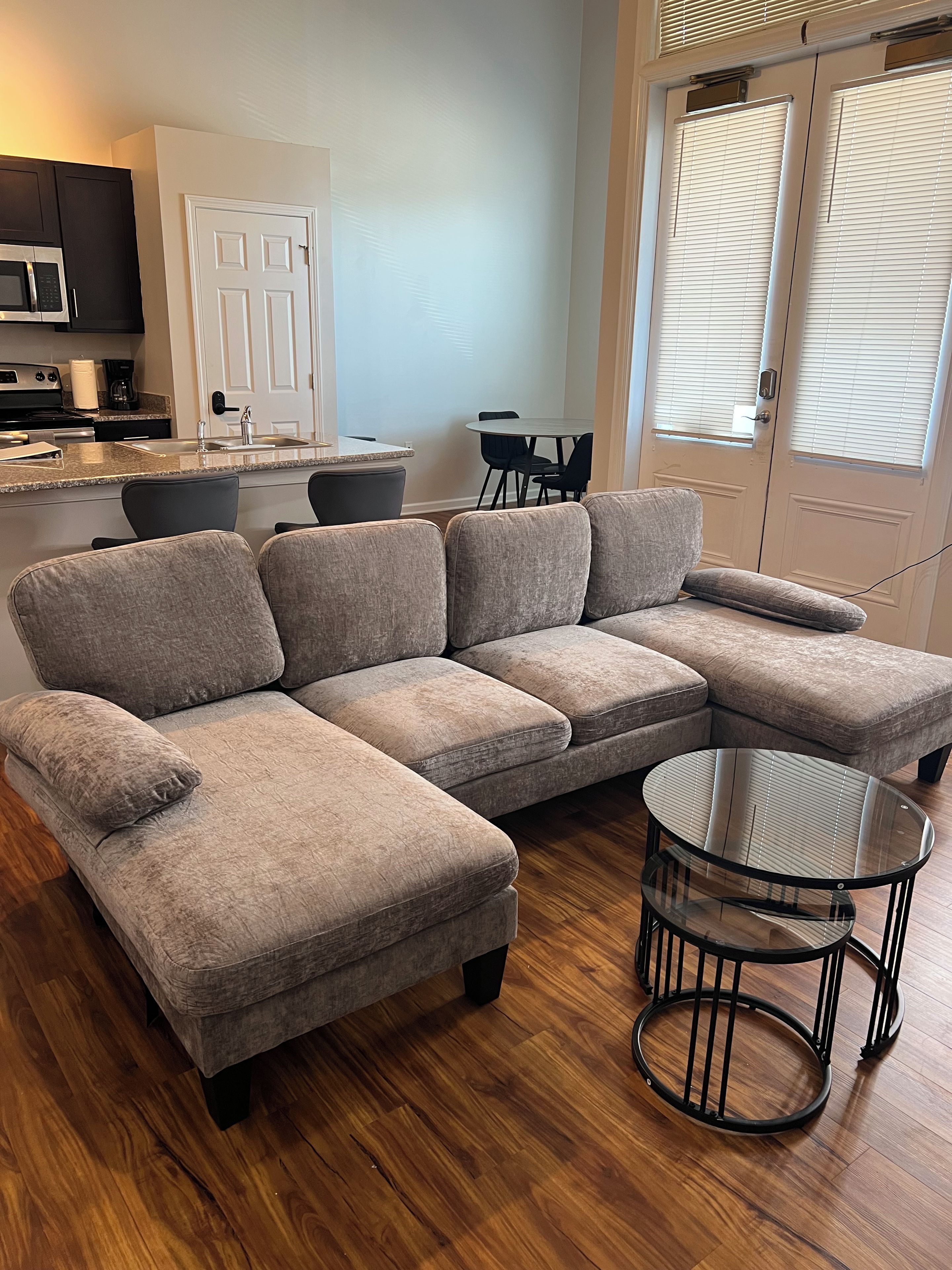 Ein modernes Wohnzimmer mit einem Ecksofa, einem Glastisch und einer Küchenzeile im Hintergrund.
