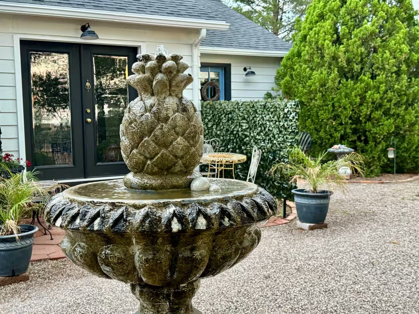 A stone fountain with a pineapple top, surrounded by gravel, with a house and greenery in the background.