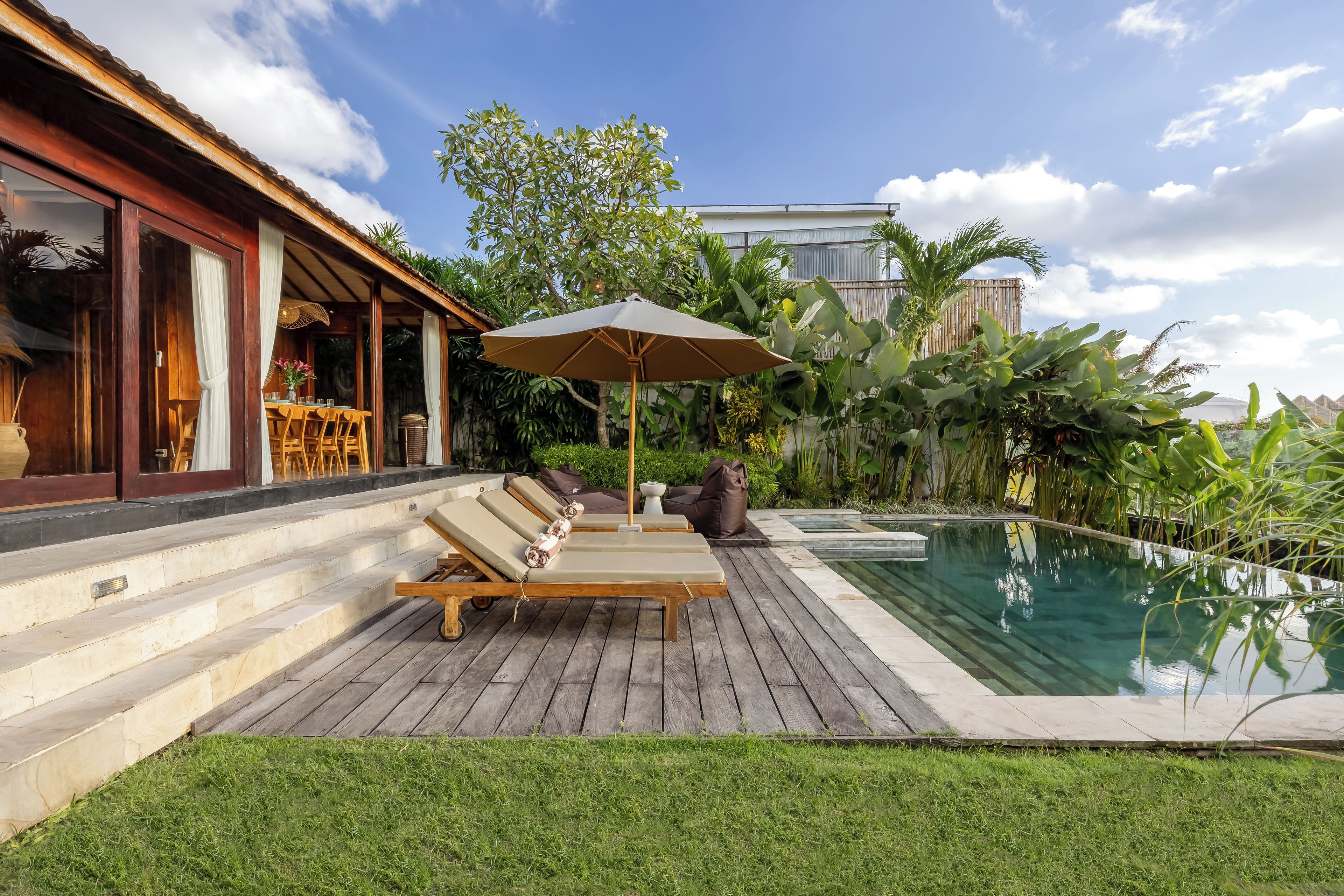 Zona junto a la piscina con sillones de descanso y una sombrilla, rodeada de exuberante vegetación y un cielo despejado.