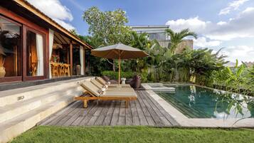 Zona junto a la piscina con sillones de descanso y una sombrilla, rodeada de exuberante vegetación y un cielo despejado.