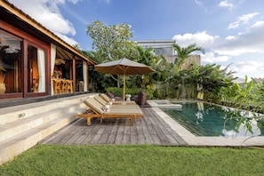 Zona junto a la piscina con sillones de descanso y una sombrilla, rodeada de exuberante vegetación y un cielo despejado.
