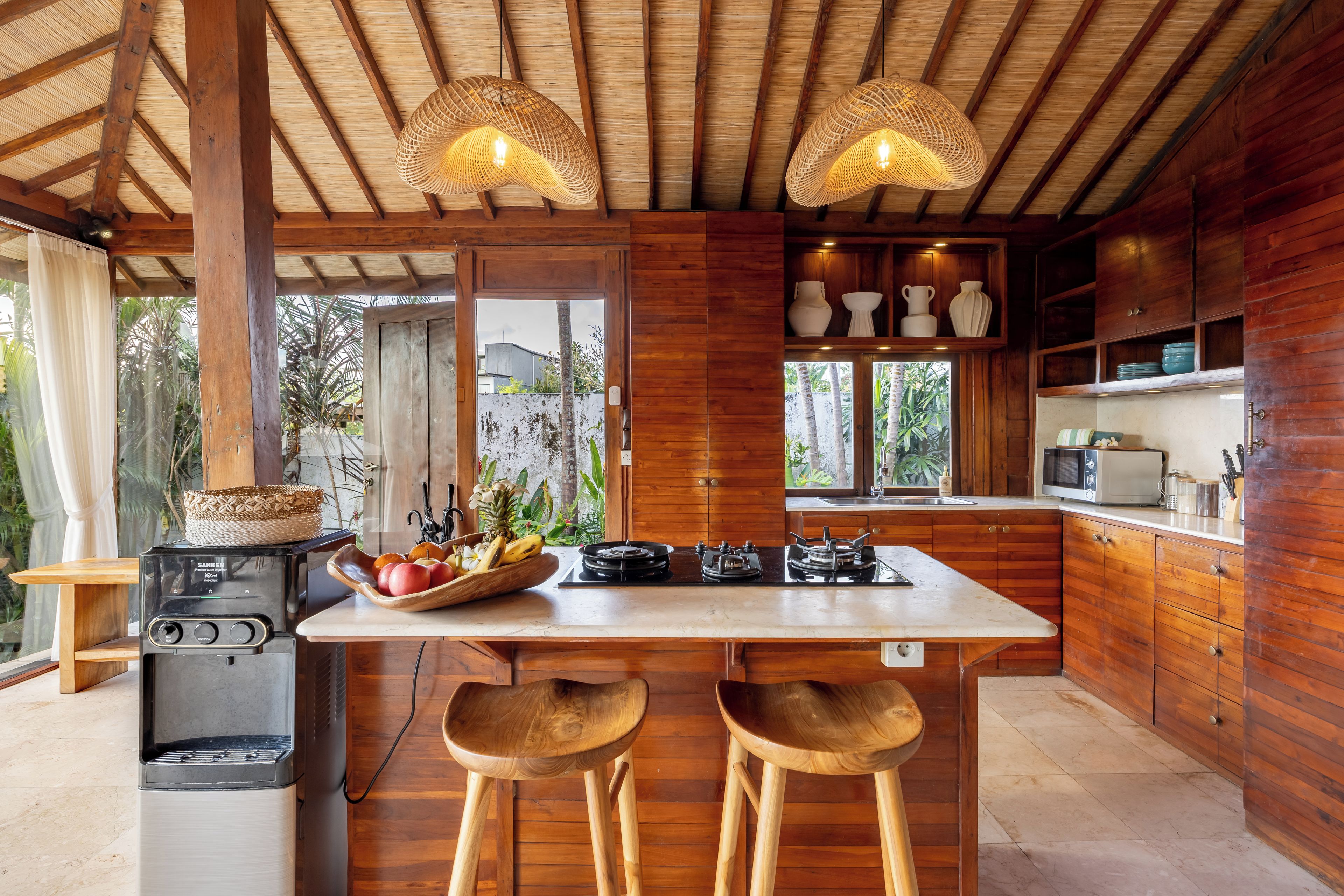 Una cocina con armarios de madera, una isla central con taburetes, una estufa a gas y lámparas colgantes tejidas.